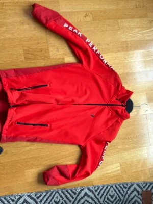Röd jacka från Peak Performance - Snygg röd jacka från Peak Performance med dragkedja framtill och två fickor med dragkedja. Jackan har logga på bröstet och text längs ärmen. Perfekt för en sportig look och passar till många tillfällen.