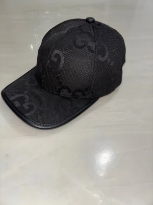 Svart keps från Gucci med GG-mönster - Snygg svart keps från Gucci med diskret GG-monogram över hela kepsen. Kepsen har böjd skärm med läderdetalj och justerbar rem baktill. Tillverkad i textilmaterial med exklusiv känsla, perfekt för en trendig streetstyle-look.