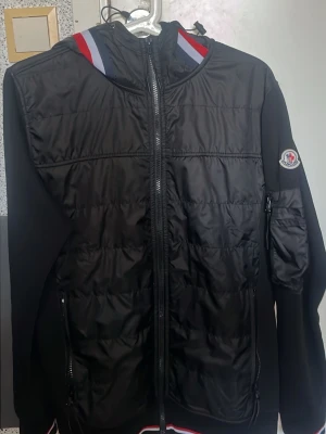 Svart pufferjacka från Moncler - (FINNS ALLT KLÄDER INNE BARA SKRIV PRIVAT ALLT FINNS )Svart pufferjacka från Moncler med dragkedja framtill, vadderad front och ärmmärke. Jackan har huva med röd, vit och blå detalj samt fickor med dragkedja. Perfekt för kalla dagar plus varma  dagar funkar året runt skyddar mot vind och kalla väder och däremot är bra för bra väder där man bara har en t Shirt under, och snygg streetstil.