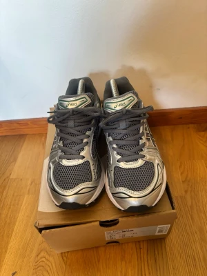 ASICS Gel-Kayano 14 silver/grå - Snygga ASICS Gel-Kayano 14 sneakers i silver och grå med meshpaneler och svarta detaljer. Skorna har en sportig retrostil, robust sula och klassisk snörning. Perfekta för dig som gillar streetwear och vill ha bekväma sneakers med cool design.