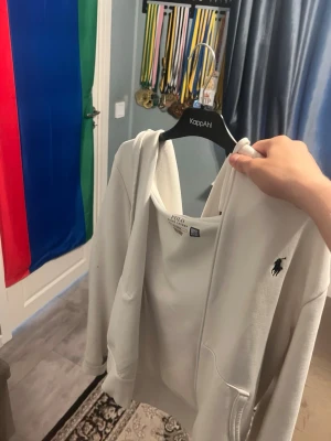 Vit hoodie från Polo Ralph Lauren - Vit hoodie från Polo Ralph Lauren med klassisk huva och dragkedja framtill. Ikonisk svart logotyp broderad på bröstet. Mjuk bomullskänsla och ribbade muddar vid ärmslut och nederkant. Perfekt för en avslappnad och stilren look.