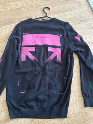 Svart Off-White tröja med rosa tryck - Säljer en svart tröja från Off-White med ikoniska rosa och svarta pilar på ryggen och rosa ränder på ärmarna. Tröjan har rund hals, långa ärmar och är gjord i ett mjukt, tunt material. Off-White-logga på bröstet. Perfekt för dig som gillar streetwear och statement-plagg.