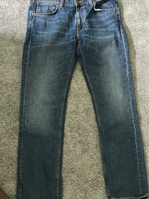 Blå jeans från Nudie Jeans Co - Säljer ett par klassiska blå jeans från Nudie Jeans Co i storlek 34/32. Jeansen har raka ben, fem fickor och snygga slitningar framtill. Tillverkade i slitstark denim med orangea sömmar och en tidlös look.