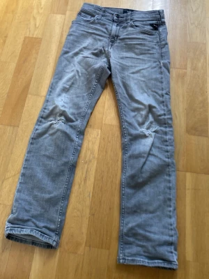 Grå slitna replay jeans - Säljer ett par grå jeans med raka ben och slitningar på knäna. Jeansen har klassisk femficksdesign och är tillverkade i bomullsdenim. Perfekt för dig som gillar en avslappnad och trendig look.