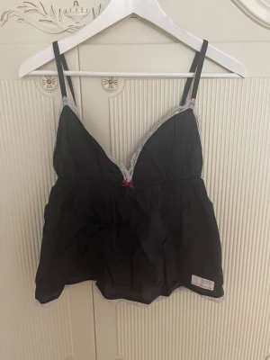 Svart linne från Odd Molly i storlek S i mycket fint skick  - Säljer linne från Odd Molly med tunna axelband och v-ringning i storlek S nyskick. Kanterna är dekorerade med vit spets och framtill finns en liten rosa rosett. Materialet är tunt och lätt, perfekt för lager-på-lager. Odd Molly-logga på ryggen.