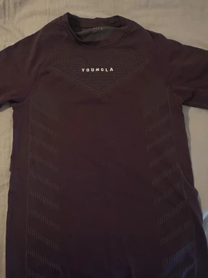 Mörklila compression från YoungLA - Snygg mörklila compression från YoungLA med korta ärmar och diskret mönster längs sidorna. Toppen har en tight passform och logga framtill. Perfekt för dig som vill ha en sportig och stilren look på gymmet.