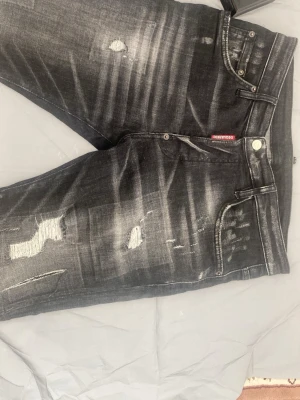 Svarta slitna jeans från Dsquared2 - Säljer ett par svarta jeans från Dsquared2 med kraftiga slitningar och distressed detaljer. Jeansen har fem fickor, silverfärgade nitar och en röd Dsquared2-tag vid fickan. Materialet är denim med stretch för skön passform.