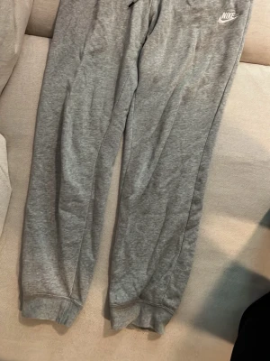 Grå mjukisbyxor från Nike - Säljer ett par grå mjukisbyxor från Nike med klassisk logga broderad på höften. Byxorna har resår i midjan med snörning och ribbade muddar vid bensluten. Perfekta för chill eller träning, riktigt sköna och stilrena.