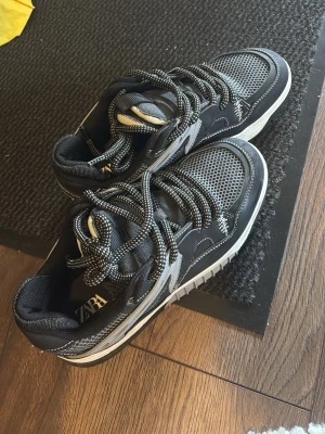 Svarta sneakers från Zara - Snygga svarta sneakers från Zara med vita och grå detaljer. Ovandelen har mesh och läderimitation, och skorna har tjocka, mönstrade snören. Sulan är vit med svarta inslag och ger en sportig vibe. Perfekta för dig som gillar streetstyle.