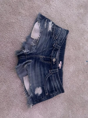 Blå slitna jeansshorts med fransar - Säljer ett par blå jeansshorts med slitningar och fransiga kanter. Shortsen har klassisk femficksmodell, dekorativa sömmar och nitar på bakfickorna. Perfekta för en avslappnad och trendig look.