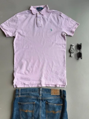 Ljusrosa pikétröja från Polo Ralph Lauren - Säljer en ljusrosa pikétröja från Polo Ralph Lauren med klassisk krage och två knappar. Tröjan har den ikoniska broderade loggan i grönt på bröstet och är gjord i mjuk bomull. Perfekt för en avslappnad men stilren look.