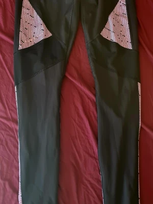 Svarta leggings med mönstrade detaljer - Snygga svarta leggings med lila och svarta geometriska mönster på sidorna och nedre delen av benen. Tight passform och stretchigt material, perfekta för träning eller en sportig look. Hög midja och paneler som ger en cool kontrast.