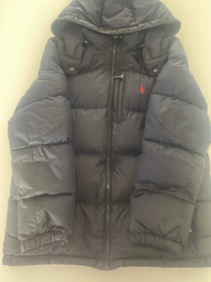Mörkblå pufferjacka från Polo Ralph Lauren - Snygg mörkblå pufferjacka från Polo Ralph Lauren med huva och dragkedja framtill. Jackan har en bröstficka med dragkedja och den klassiska röda Polo-loggan broderad på bröstet. Perfekt för kalla dagar och riktigt skön att bära.