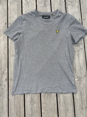 Grå t-shirt från Lyle & Scott - Snygg grå t-shirt från Lyle & Scott med klassisk gul logga på bröstet. T-shirten har rund hals, korta ärmar och är gjord i mjuk bomull. Perfekt basplagg till garderoben. Storlek M