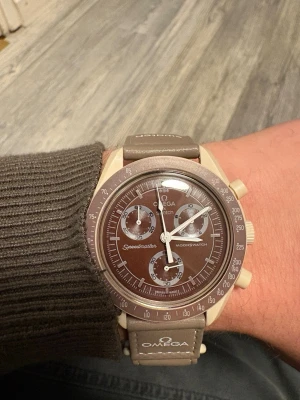 Omega x Swatch - Snygg brun Omega x Swatch Speedmaster Moonswatch med beige detaljer och matchande brunt armband. Klockan har flera urtavlor och tydliga vita markeringar. Armbandet är i syntetmaterial och har Omega-loggan tryckt på sidan.