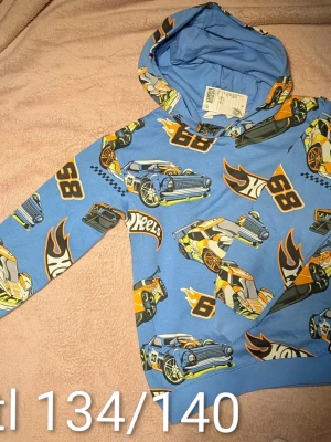 Ny Blå Hot Wheels hoodie med bilar - Cool blå hoodie från Hot Wheels med färgglada bilmotiv, flames och nummertryck. Tröjan har huva och ribbade mudd. Materialet är mjukt och bekvämt. Finns i Holmsund, skickas mot porto.