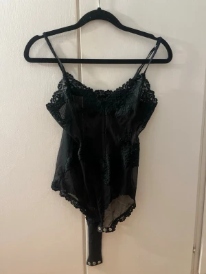 Svart spetsbody från Intimissimi  - Säljer en svart body från Intimissimi med tunna axelband och vacker spetsdetalj upptill och längs kanterna. Bodyn är i spets, har tryckknappar i grenen och ett transparent, elegant utseende. Perfekt för dig som gillar stilrena underkläder med en twist.
