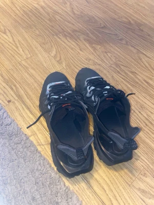 Nike svarta sneakers med reflexdetaljer - Svarta Nike sneakers med grå och vita reflexdetaljer, svarta snören och robust sula. Swoosh-logga på sidan och orangea accenter vid snörningen. Ovandel i mesh och syntetmaterial, låg modell med modern design.