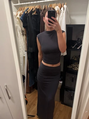 Mörkgrått tvådelat set – crop top & maxikjol | Gina Tricot | Topp S / Kjol M  - Snyggt och minimalistiskt set från Gina Tricot i en mörkgrå/antracit färg. Crop topen har polokrage och korta ärmar med lätt paddad axeldetalj som ger en snygg silhuett. Maxikjolen är tajt och stretchig med ruching längs sidan. Tillsammans ger de ett clean och elegant intryck som passar både vardag och fest.  Knappt använt, i nyskick. Topp strl S, kjol strl M – passar perfekt ihop på en S.