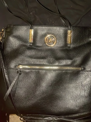 Svart handväska från Michael Kors - Snygg svart handväska från Michael Kors i strukturerat konstläder med guldfärgade detaljer och logotyp framtill. Väskan har dubbla handtag, en dragkedjeficka på framsidan och stilren design som passar till många outfits.knappt använd 