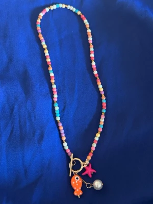 Färgglatt pärlhalsband med berlocker - Ett halsband med små färgglada pärlor i rosa, blått, gult och vitt. Smycket har tre berlocker: en orange fisk, en rosa sjöstjärna och en vit pärla. Halsbandet har ett guldfärgat lås och ger en lekfull och trendig vibe.
