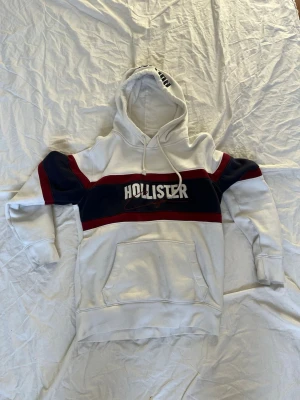 Vit hoodie från Hollister med ränder - Snygg vit hoodie från Hollister med marinblå och röda ränder över bröstet samt stor logga framtill. Hoodien har huva med dragsko och en stor magficka. Perfekt för en avslappnad och trendig stil.