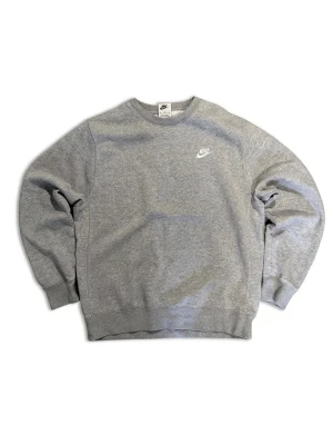 Grå sweatshirt från Nike - Klassisk grå sweatshirt från Nike med broderad vit logga på bröstet. Tröjan har rund halsringning, ribbade muddar vid ärmslut och nederkant. Perfekt för en avslappnad och sportig stil.