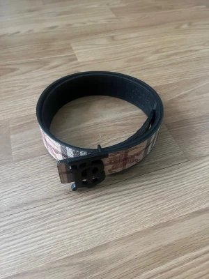 Rutig Burberry-bälte med TB-spänne - Snyggt bälte från Burberry med klassiskt rutigt mönster i beige, brun och röd. Bältet har ett svart TB-monogram som spänne och är gjort i skinn. Perfekt accessoar för att lyfta din outfit.
