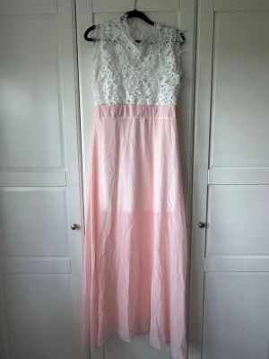 Långklänning med spets och rosa kjol - Romantisk långklänning med vit spets upptill och ljusrosa, flowy kjol. Ärmlös modell och detaljerad spets på överdelen. Perfekt för student eller sommarens speciella tillfällen. Strl S