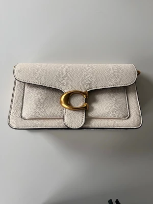 Vit kuvertväska från Coach - Stilren vit kuvertväska från Coach i strukturerat läder med guldfärgat C-spänne framtill. Väskan har en rektangulär form och minimalistisk design, perfekt för att lyfta din outfit. Klassisk och tidlös look med exklusiva detaljer.