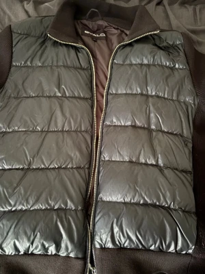 Svart dunväst från Bvaldi - Snygg svart cardigan från Bvaldi med quiltad design och dragkedja framtill. Cardigan har ribbade detaljer vid krage och sidor samt en ficka med knapp och Bvaldi-logga på ärmen. 