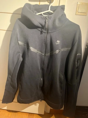 Svart Nike hoodie med dragkedja - Snygg svart hoodie från Nike med hel dragkedja framtill och huva. Har en diskret logga på bröstet, tejpade detaljer över bröstet och en praktisk ficka med dragkedja på ärmen. Tillverkad i mjukt material som känns bekvämt och sportigt.