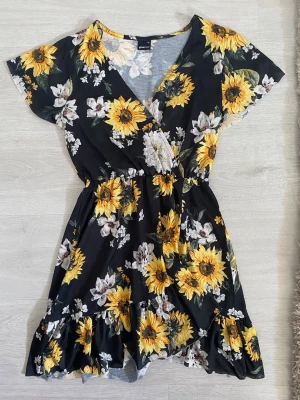Svart blommig omlottklänning - Kort svart omlottklänning från Gina Tricot med solrosor och vita blommor. Klänningen har volangkant nertill, v-ringning och korta ärmar. Perfekt för sommarens alla tillfällen och har en lätt och luftig känsla.