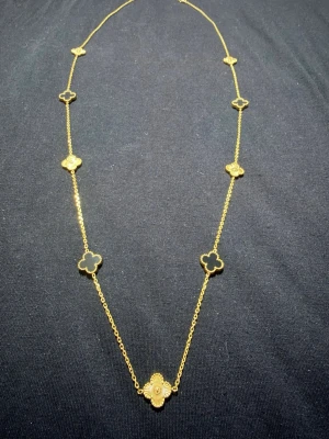 Guldhalsband med klöverhängen - Snyggt halsband i guldton med flera fyrklöverformade hängen längs kedjan. Hängena har en elegant och stilren design, vissa med svart detalj och andra i helguld. Perfekt accessoar för att lyfta din outfit.