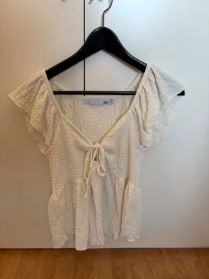 Vit blus med volang och knyt - Söt vit blus med volangärmar och knyt framtill. Blusen har en lätt rynkad struktur och v-ringning. Perfekt för en somrig look och passar till både jeans och kjol.