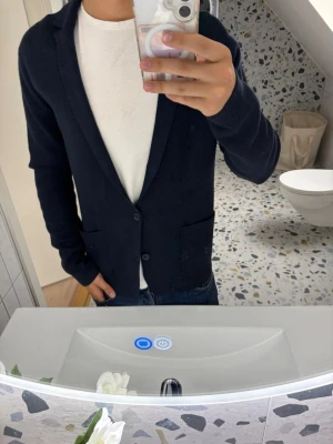 Mörkblå stickad kavaj - Stilren mörkblå stickad kavaj med två knappar framtill och två praktiska fickor. Klassisk krage och lång ärm, perfekt att slänga över en t-shirt för en clean look. Materialet ser ut att vara mjukt och stickat, vilket ger en avslappnad men ändå uppklädd känsla.