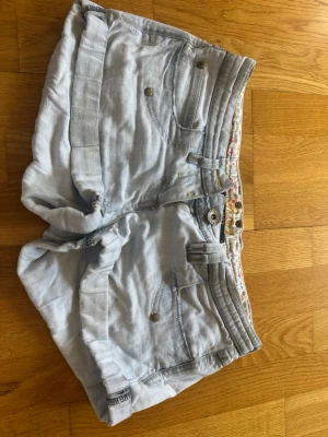 Låg midja shorts - Säljer ett par ljusblå denimshorts med hög midja och flera dekorativa sömmar i midjan. Shortsen har fickor både fram och bak samt bälteshällor. Perfekta för sommaren och ger en avslappnad vibe.