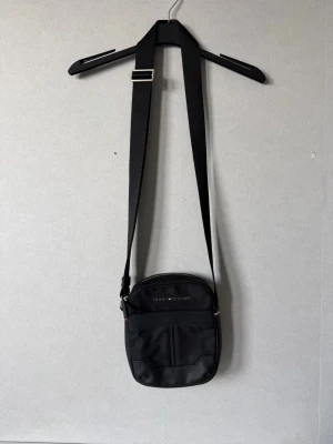 Svart axelväska Tommy Hilfiger - Snygg svart axelväska från Tommy Hilfiger med justerbar axelrem och minimalistisk design. Väskan har ett ytterfack framtill och diskret logga upptill. Tillverkad i slitstarkt syntetmaterial. Har använts 2 gånger. 