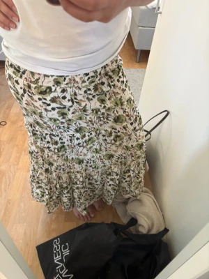 Blommig långkjol  - Säljer en långkjol med blommigt mönster i vitt, grönt och inslag av rosa. Kjolen har volangdetalj nedtill och är tillverkad i ett lätt, luftigt material som passar perfekt för varmare dagar. Resår i midjan för skön passform. Helt oanvänd!