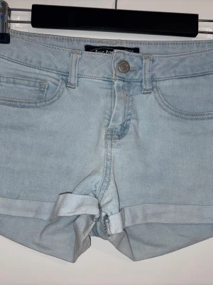 Ljusblå denimshorts från JayJays - Säljer ett par ljusblå denimshorts från JayJays med klassisk femficksdesign och uppvikta benslut. Shortsen har normal passform, bälteshällor och stängs med knapp och dragkedja. Perfekta för varma dagar och enkel att matcha med olika toppar.