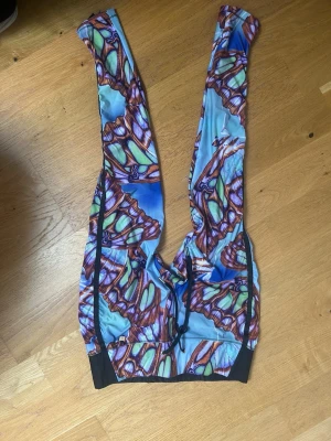 Tights med fjärilsmönster Power Woman - Säljer ett par färgglada cykelbyxor från Power Woman med unikt fjärilsmönster i blått, grönt och brunt. Byxorna har breda axelband och svart resår i midjan för extra komfort. Perfekta för cykling och träning.