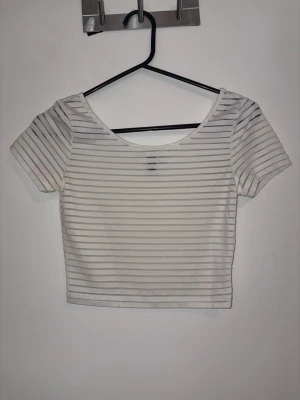Vit randig croppad topp från Divided - Säljer en vit croppad topp från Divided med tunna horisontella ränder. Toppen har korta ärmar och en rundad halsringning. Perfekt till jeans eller kjol för en enkel och fräsch look.