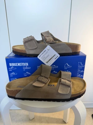 Birkenstock Arizona taupe sandaler - Säljer ett par Birkenstock Arizona sandaler i färgen taupe. De har två justerbara remmar med metallspännen och mjuk fotbädd i mocka. Sulan är i kork och gummi för extra komfort. Perfekta för dig som gillar klassisk och stilren design.