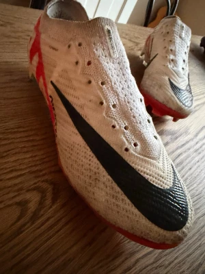 Nike vita mercurials elite fotbollsskor med svart swoosh - Säljer ett par vita Nike fotbollsskor med stor svart swoosh på ovansidan och röda detaljer på sidan och sulan. Skorna har en strumpliknande passform och är tillverkade i ett stickat material för bra komfort och bollkontroll. Perfekta för dig som vill ha stilrena och lätta skor på planen.