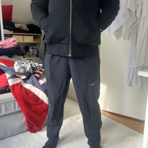 Svart hoodie med dragkedja från Nike - Snygg svart hoodie från Nike med dragkedja framtill och två fickor på sidorna. Klassisk design med huva och ribbade muddar vid ärmslut och nederkant. Perfekt för en avslappnad och sportig stil. Nike-logga på bröstet för en clean look.
