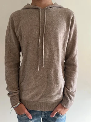 Beige hoodie i mjukt material - Säljer en beige hoodie med huva och dragsnören. Hoodien har en klassisk känguruficka framtill och ribbade muddar vid ärmslut och nederkant. Tillverkad i ett mjukt, stickat material som känns skönt mot huden. Perfekt för en avslappnad och stilren look.