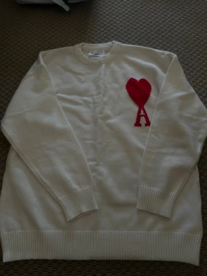 Sweater Ami Paris Ami de Coeur Off-White - Vit stickad tröja från Ami Paris med rund halsringning och ribbade muddar. På bröstet finns ett rött hjärta med bokstaven 'A' i samma färg, vilket är signaturdetaljen för märket. Perfekt för dig som gillar stilrena och trendiga plagg.