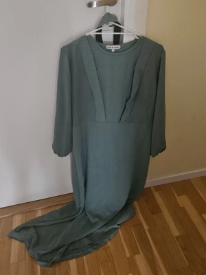 Lång grön klänning från Turkiet  - Elegant långklänning i dämpad grön nyans från Turkiet. Klänningen har långa ärmar och en stilren, minimalistisk design med markerade sömmar framtill. Perfekt för dig som gillar en enkel men sofistikerad look. ANVÄNDTS 2 GÅNGER.