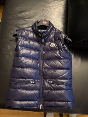 Mörkblå dunväst från Moncler - Snygg mörkblå dunväst från Moncler med hög krage och dragkedja framtill. Västen har två fickor med dragkedja och det klassiska Moncler-logotypen på bröstet. Perfekt för lager på lager och ger en clean look till din outfit.