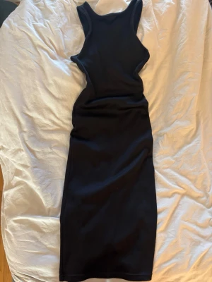 Svart ribbad långklänning - Säljer en svart ribbad långklänning med ärmlös design och rund halsringning. Klänningen har en slits bak för extra rörelsefrihet och en tight passform som framhäver figuren. Perfekt för dig som gillar stilrena och enkla plagg.